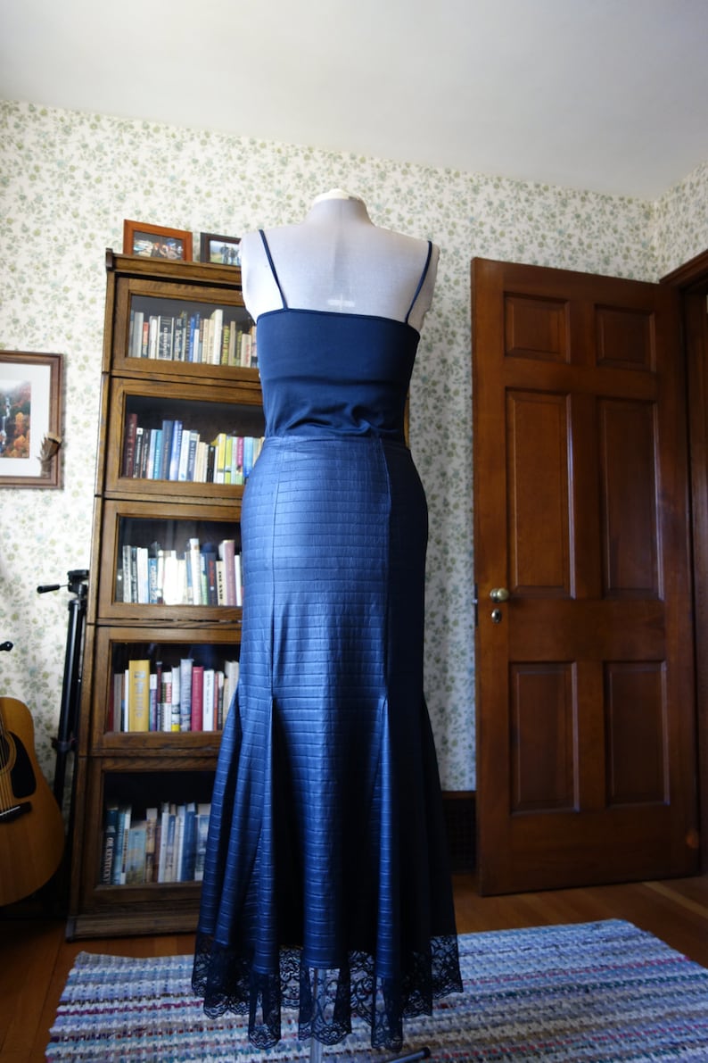 May include: A dark blue mermaid-style skirt with a black lace trim. The skirt is on a mannequin and is displayed in front of a bookcase and a wooden door.