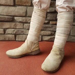 May include: A pair of white suede boots with a unique design featuring multiple straps. The boots are worn and have a distressed look.