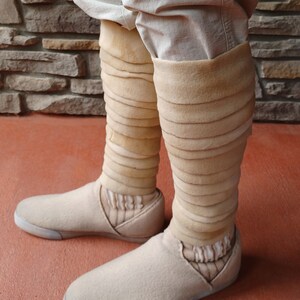 May include: A pair of beige felt shoes with a white sole. The shoes have a unique design with multiple layers of beige fabric wrapped around the ankles.