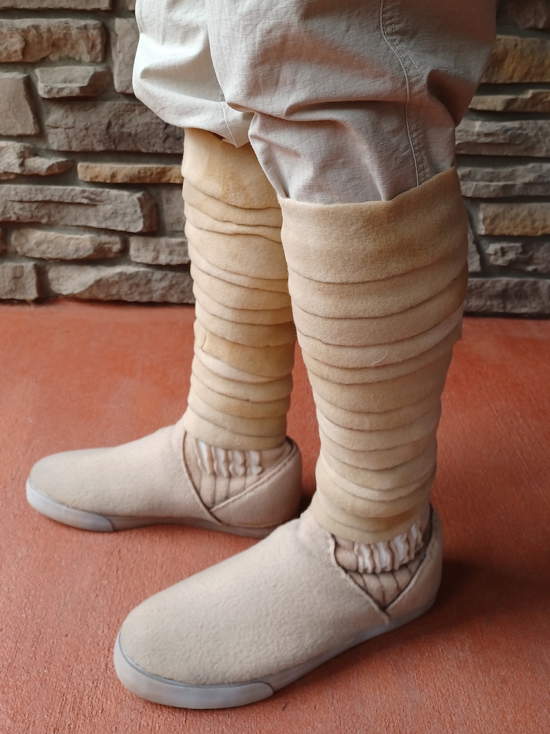 May include: A pair of beige felt shoes with a white sole. The shoes have a unique design with multiple layers of beige fabric wrapped around the ankles.