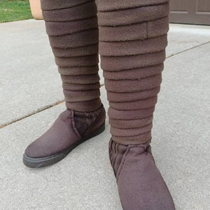 May include: A pair of brown, knee-high boots with a layered, folded design. The boots are made of a soft, fuzzy material and have a black sole.