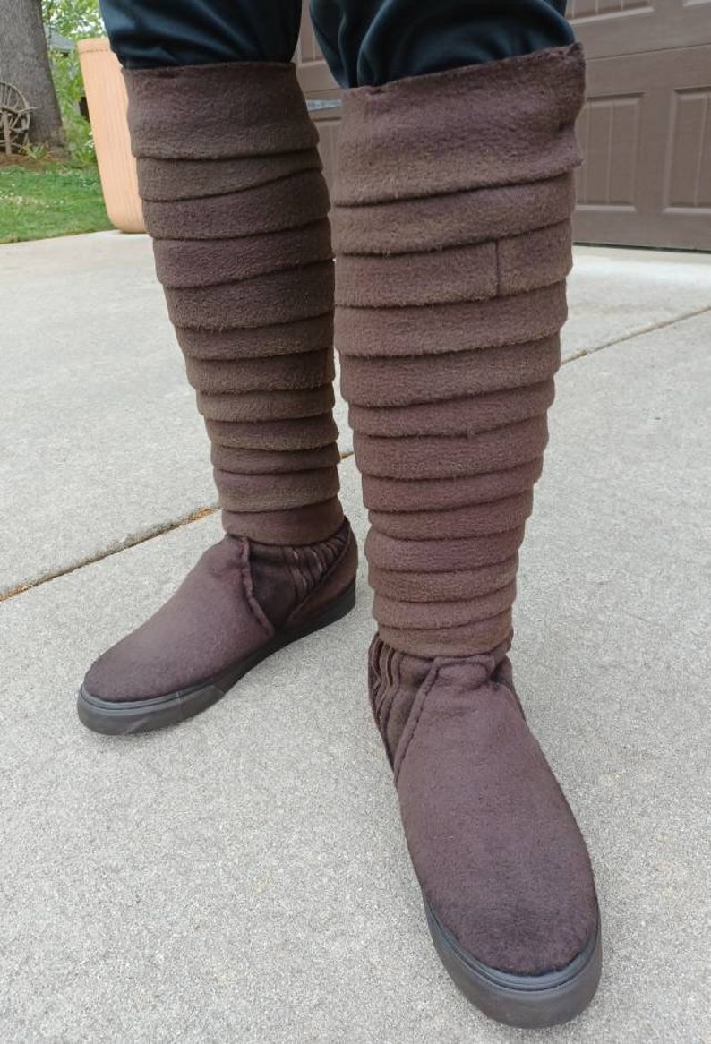 May include: A pair of brown, knee-high boots with a layered, folded design. The boots are made of a soft, fuzzy material and have a black sole.