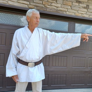May include: A man wearing a white robe with a brown belt and a silver buckle. He is standing in front of a brown wooden door with a window above it. He is pointing to the right with his right hand.