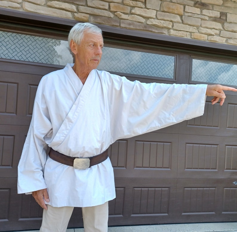 May include: A man wearing a white robe with a brown belt and a silver buckle. He is standing in front of a brown wooden door with a window above it. He is pointing to the right with his right hand.