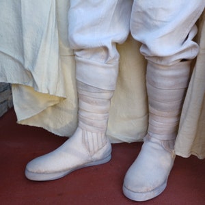 May include: A pair of white, canvas boots with a thick, layered, white fabric cuff. The boots are worn and have a distressed look.