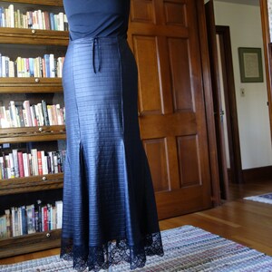 May include: A black skirt with a lace trim and a textured, quilted design. The skirt is on a mannequin and is displayed on a wooden floor with a patterned rug.
