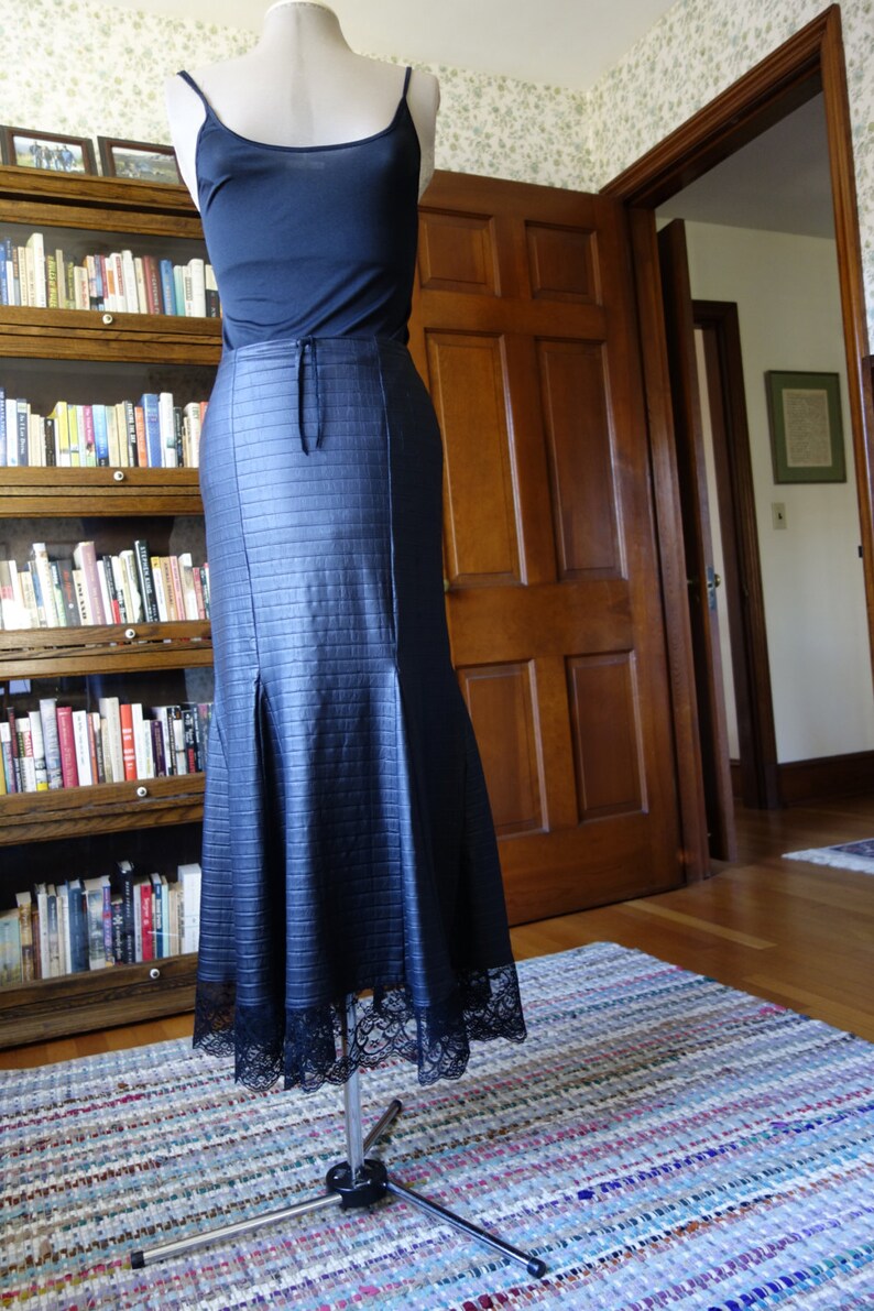 May include: A black skirt with a lace trim and a textured, quilted design. The skirt is on a mannequin and is displayed on a wooden floor with a patterned rug.