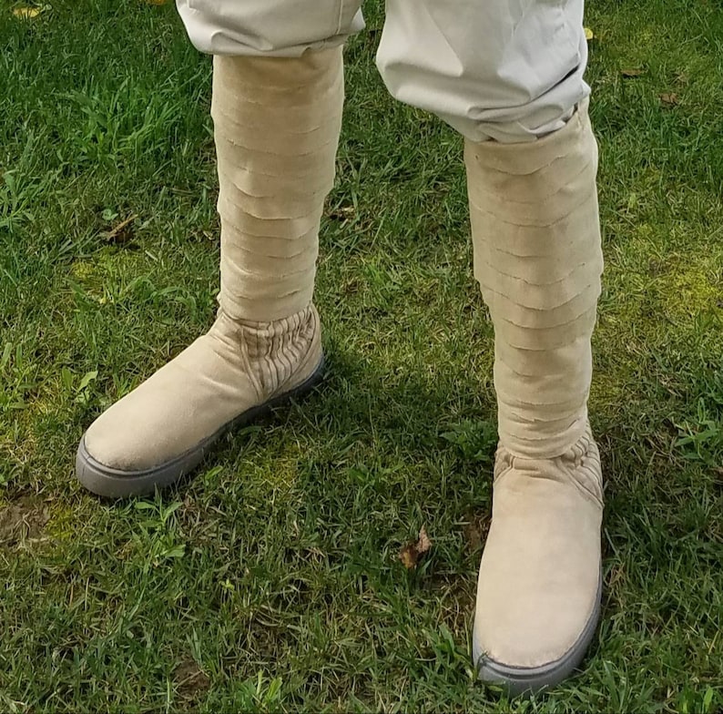 May include: A pair of beige boots with a unique layered design. The boots are made of a soft, textured material and have a rounded toe.