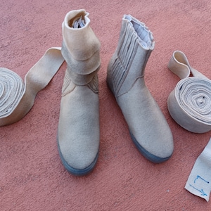 May include: Two pairs of beige felt boots with a rolled cuff. The boots are on a pink surface. There are two rolls of beige felt fabric next to the boots.