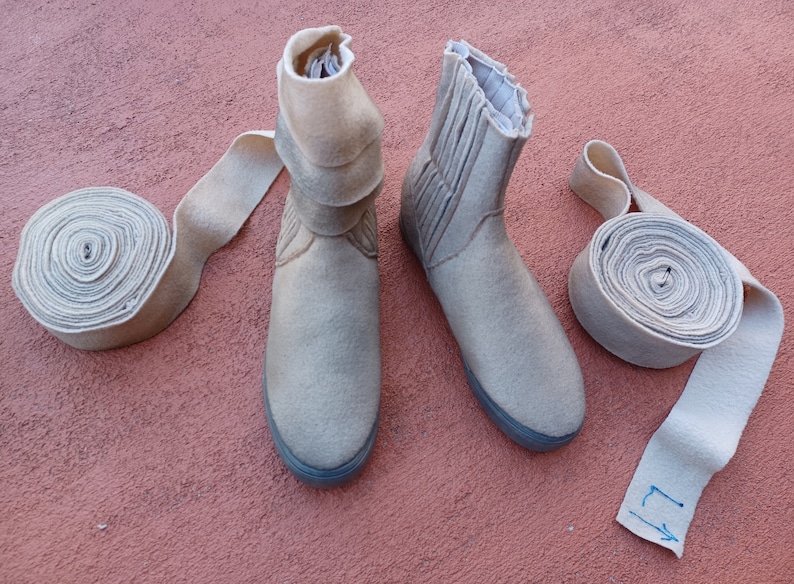May include: Two pairs of beige felt boots with a rolled cuff. The boots are on a pink surface. There are two rolls of beige felt fabric next to the boots.