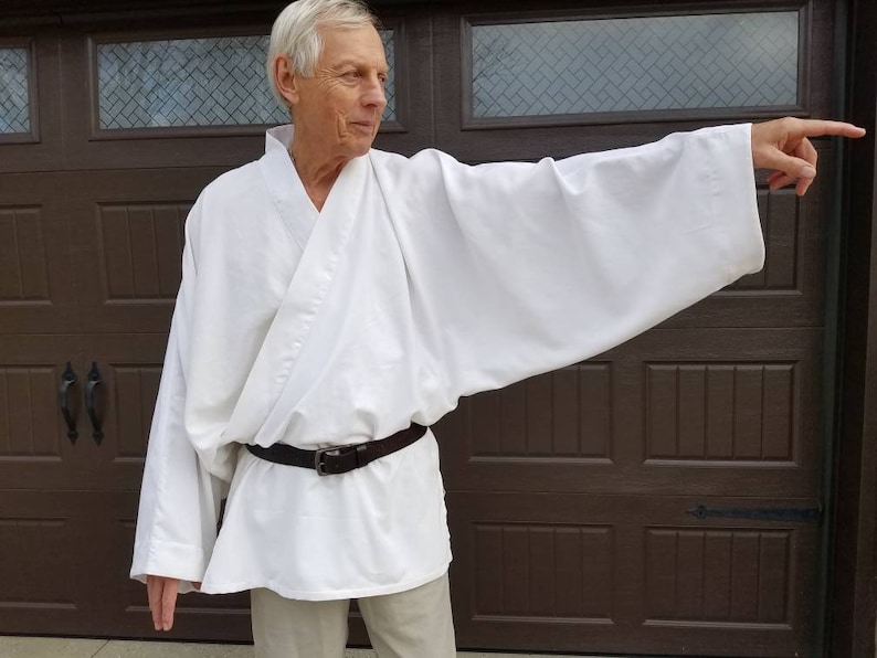 May include: A white robe with long sleeves and a brown belt. The robe is worn by a person standing in front of a brown garage door.