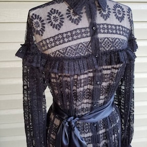 May include: A black lace dress with a button-up front, long sleeves, and a ruffled collar. The dress has a black satin sash tied at the waist.