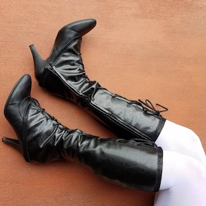 May include: A pair of black leather knee-high boots with laces. The boots have a pointed toe and a high heel. The boots are worn with white tights.