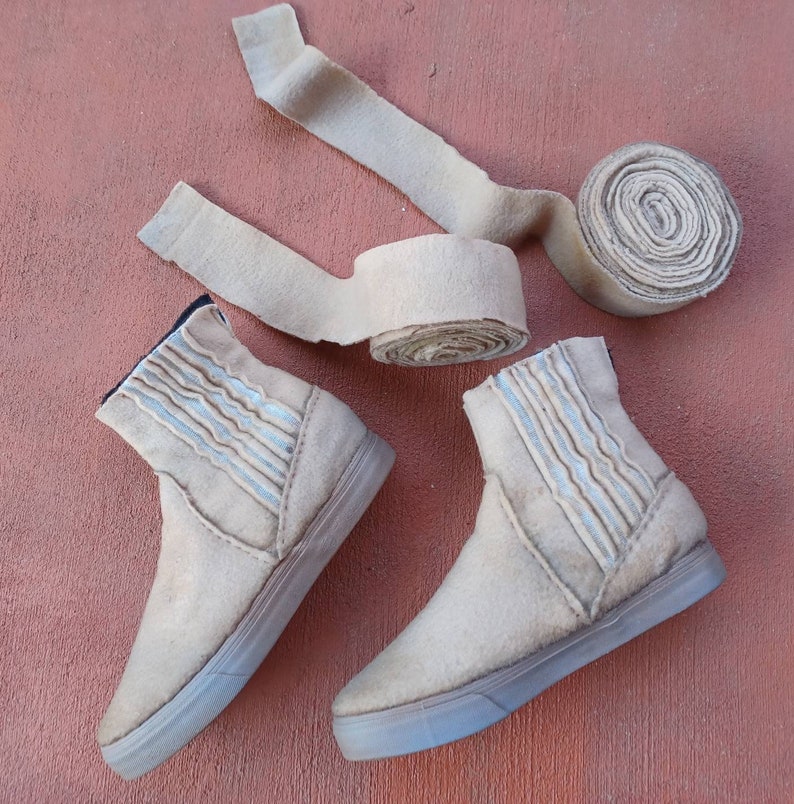 May include: Two pairs of beige felt ankle boots with white stitching. The boots have a rounded toe and a flat sole. The boots are on a brown surface.