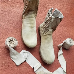 May include: Two pairs of beige suede boots with a zipper on the side. The boots are on a red surface with two beige suede straps.
