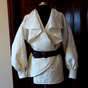May include: Cream-colored quilted jacket with a wrap-style design and a wide collar. The jacket features intricate floral patterns and is cinched at the waist with a dark brown belt. The sleeves are long and gathered at the cuffs.
