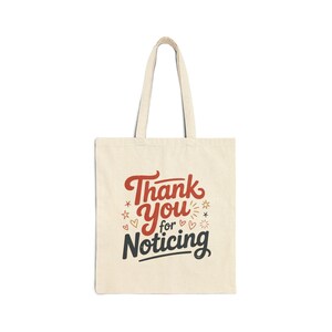 May include: A cream-coloured tote bag featuring the text "Thank You for Noticing" in red, black, and cream. The design includes stars and hearts. The bag has two cream-coloured handles.