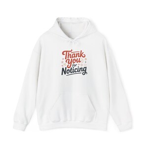 Puede incluir: Sudadera con capucha blanca con bolsillo delantero y cordón. La parte delantera de la sudadera presenta el texto "Thank You for Noticing" en letras rojas y azul marino con estrellas y corazones decorativos.