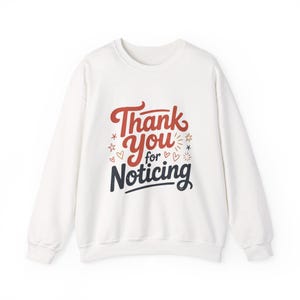 Puede incluir: Sudadera blanca de cuello redondo con la frase "Thank You for Noticing" en letras rojas y azul marino. El diseño incluye estrellas y corazones. Una prenda superior informal y cómoda.