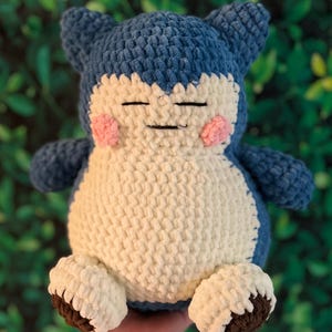 May include: A handmade crochet plush toy resembling a cartoon character. It features a blue head and limbs, a cream body, and pink cheeks. The feet are cream with brown soles. The toy is approximately 25 cm tall.