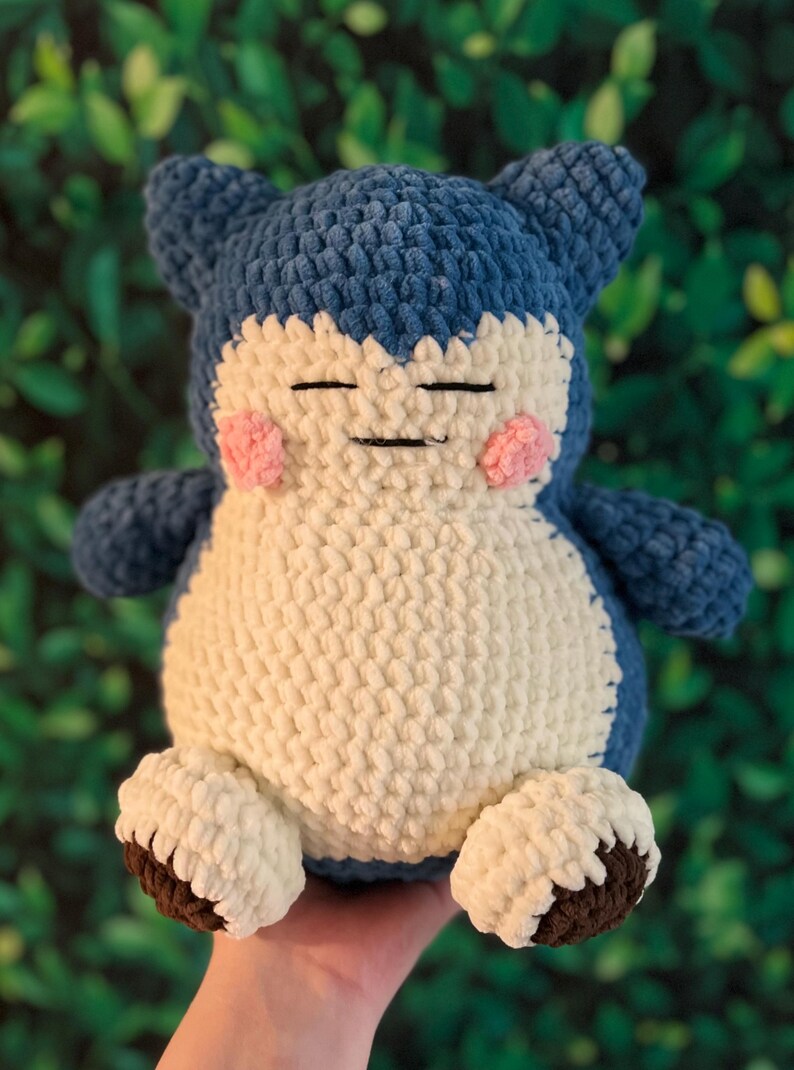 May include: A handmade crochet plush toy resembling a cartoon character. It features a blue head and limbs, a cream body, and pink cheeks. The feet are cream with brown soles. The toy is approximately 25 cm tall.