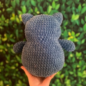 May include: A blue crochet teddy bear with small ears and arms. The soft toy is made of a textured yarn and is held in a hand against a green leafy background. The bear is a cute, cuddly item, perfect for a child's bedroom.