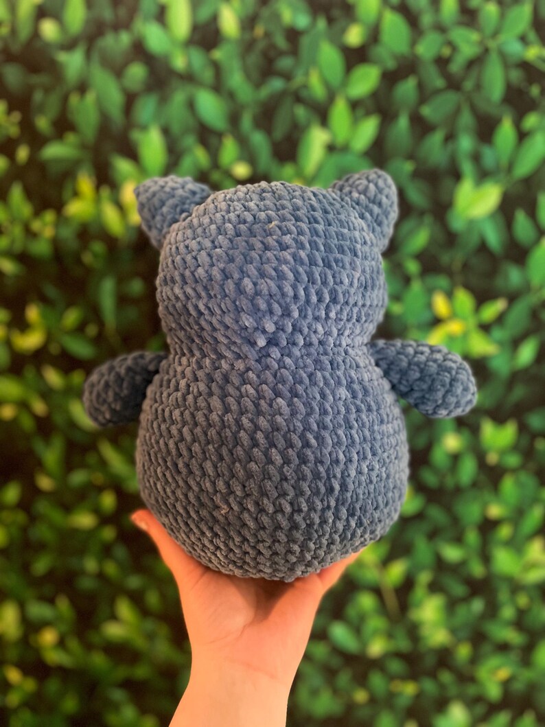 May include: A blue crochet teddy bear with small ears and arms. The soft toy is made of a textured yarn and is held in a hand against a green leafy background. The bear is a cute, cuddly item, perfect for a child's bedroom.