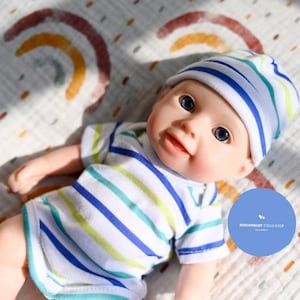 Può includere: Una bambola realistica con un body a righe blu, verdi e bianche e un berretto abbinato. La bambola ha gli occhi azzurri e un'espressione sorridente. La bambola è sdraiata su una coperta bianca con un disegno arcobaleno.