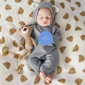 以下が含まれることがあります： グレーのクマをテーマにした衣装を着た赤ちゃん人形。お揃いのフードと耳が付いており、茶色のクマのプリントが入った白い毛布の上に横たわっています。小さなぬいぐるみと金色の pacifier クリップも写っています。