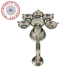May include: A silver-colored, multi-cup oil lamp with a central reservoir and six surrounding cups. The lamp has an ornate, decorative design and a handle. A circular logo with text is visible in the upper left corner.
