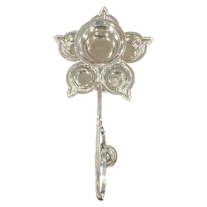 May include: A silver-colored, flower-shaped oil lamp with five circular receptacles for oil and wicks. The lamp has a long, decorative handle and a small, circular base. The design is symmetrical and detailed, suitable for religious or decorative purposes.