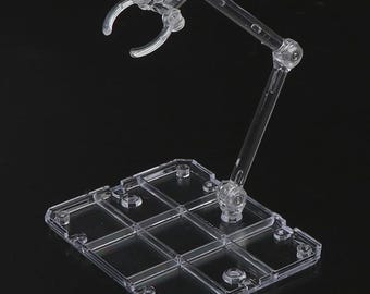 May include: A clear plastic action figure stand with a rectangular base and an adjustable arm. The stand features a claw-like grip to hold the figure and multiple joints for posing. The stand is designed to provide support and display action figures.