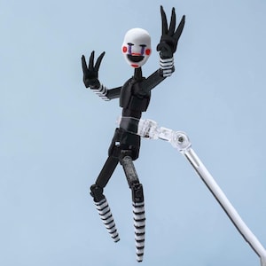 Puede incluir: Una figura de acción articulada de un personaje con máscara blanca, ojos negros, boca negra y mejillas rojas. La figura tiene un cuerpo negro con extremidades largas y delgadas, mangas y piernas a rayas blancas y negras, sobre un soporte transparente.
