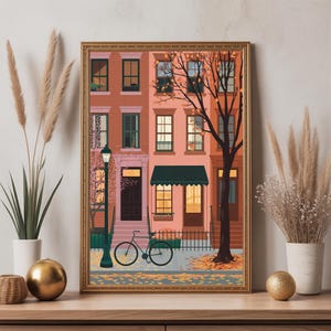 May include: A framed art print showing a street scene with brownstone buildings in peach and brown hues. A bicycle is parked on the pavement, and a bare tree stands beside the buildings. The artwork is enclosed in a gold-coloured frame.