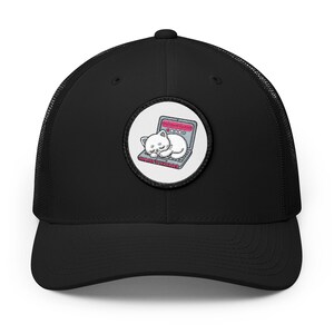 May include: A black trucker hat featuring a circular patch on the front. The patch displays a white cartoon cat sleeping on a grey laptop. The laptop screen shows a pink rating bar with stars and the text NOBODY IS MANAGER in pink letters.