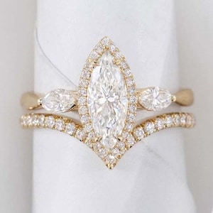 Pode incluir: Um conjunto de anéis de noivado dourados com um diamante central em corte marquise, rodeado por um halo de diamantes menores. O conjunto inclui uma banda a condizer com um design curvo e pequenos diamantes. Os anéis são exibidos numa superfície branca.