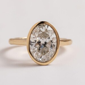 May include: A gold ring featuring an oval-cut diamond. The diamond is set in a bezel setting, and the band is a simple, polished gold. The ring is displayed on a white background, highlighting the sparkle of the diamond.