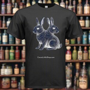 May include: Black t-shirt featuring a detailed illustration of a two-headed rabbit in shades of blue and white. The design is centered on the shirt, with the website address CocosLittleShop.com printed below the graphic. The shirt is made of a soft material.