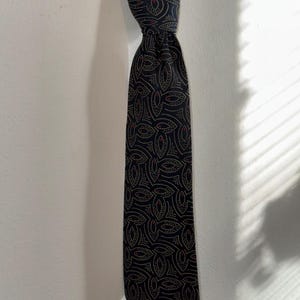 May include: A dark navy blue tie with a repeating pattern of beige and red leaf-like shapes. The tie is knotted and hangs against a white wall, with shadows from sunlight visible on the right side.