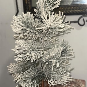 May include: A small artificial Christmas tree with green needles covered in white flocking, mimicking snow. The tree stands on a copper-coloured metal base. A festive decoration for the holiday season, it is a miniature fir tree.