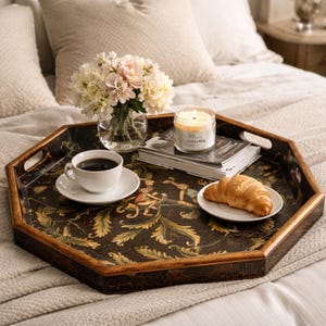 Puede incluir: Una bandeja octogonal con un diseño oscuro y ornamentado contiene una taza de café, un cruasán, una vela y un jarrón con flores. La bandeja está sobre una cama con ropa de cama de color neutro, creando una escena acogedora.