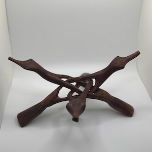 May include: A dark brown wooden tripod stand with an intricate, interwoven design. The stand has three curved legs that meet in the middle, providing a stable base. The wood has a natural, textured look, and the stand is designed to hold a round object.