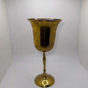 Puede incluir: Una copa dorada con un cuenco ancho grabado con motivos florales, un tallo delgado y una base redonda. La copa tiene un acabado pulido y está sobre un fondo blanco. El diseño general es elegante y vintage.