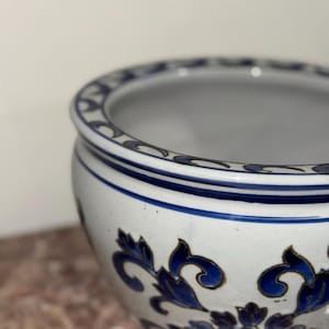 Könnte beinhalten: Eine weiße Keramikschale mit blauen Blumenmustern und Verzierungen. Die Schale hat eine weite Öffnung und eine abgerundete Form. Das blaue Blumendesign wiederholt sich um die Außenseite der Schale. Die Schale wird wahrscheinlich zu dekorativen Zwecken oder als Pflanzgefäß verwendet.