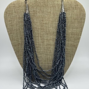 May include: A multi-strand necklace with dark gray beads. The necklace is displayed on a tan jewelry bust. The strands cascade down, creating a layered effect. The necklace has silver-toned metal accents at the top.