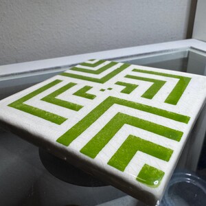 Op de afbeelding: Vierkante keramische tegel met een witte achtergrond en een geometrisch patroon in groentinten. Het ontwerp toont een reeks verbonden, hoekige lijnen. De tegel wordt waarschijnlijk gebruikt als onderzetter of decoratief element.