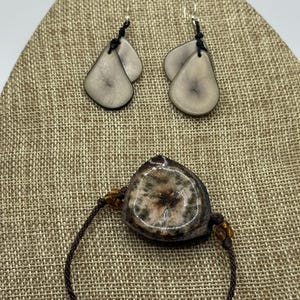 Puede incluir: Un conjunto de joyas con pulsera y pendientes. Los pendientes tienen forma de lágrima con color gris claro y detalles en negro. La pulsera tiene una piedra similar con cordón marrón y cuentas color ámbar.