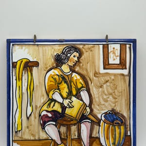 Puede incluir: Azulejo de cerámica pintado a mano con borde azul. La obra representa a una persona con ropa amarilla y burdeos sentada en un taburete, sosteniendo un libro. Una tela amarilla cuelga de un perchero de madera y se ve un cubo a rayas.