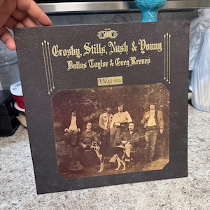 Könnte beinhalten: Ein Vintage-Vinyl-Schallplattenalbum mit dem Titel "Crosby, Stills, Nash & Young Deja Vu". Das Albumcover ist dunkelbraun mit goldfarbenen Buchstaben und einer sepiafarbenen Fotografie der Band und eines Hundes. Eine türkisfarbene Keramik-Hühnerfigur sitzt obenauf.