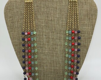 Puede incluir: Un collar de múltiples hilos con cadenas doradas y cuentas de colores. El collar presenta hilos de cuentas rojas, azules, verdes y rosas, creando un accesorio vibrante. El collar se exhibe sobre un busto de tono neutro.